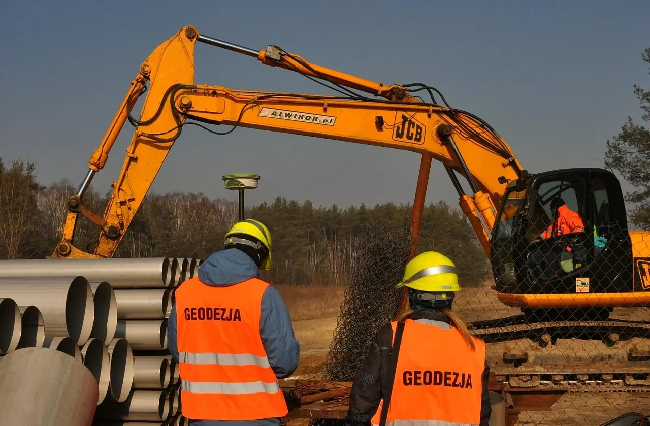 Excavator digging beside stacked pipes while two surveyors in vests watch